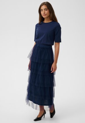 Donna in piedi che indossa una maglietta blu navy a maniche corte e una gonna maxi in tulle blu navy a strati con scarpe nere con tacchi alti su uno sfondo semplice.