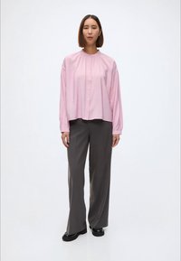 Lyserosa langermet skjorte med rynket hals, kombinert med løse grå bukser og svarte sko, satt mot en enkel hvit bakgrunn.