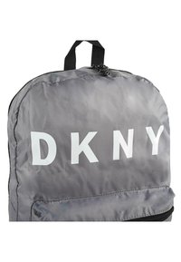 Mochila de nailon gris con un gran logo blanco de "DKNY". Tiene un asa superior, cierre de cremallera y un bolsillo frontal con una cremallera negra.