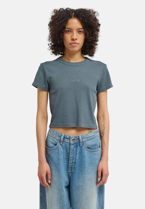Młoda kobieta o kręconych włosach, ubrana w niebieski krótki t-shirt z napisem "element" oraz luźne, wysokie niebieskie jeansy, stojąca na białym tle.