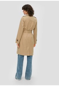 Manteau beige avec taille ceinturée, manches longues et détail dos fente, associé à un jean large bleu clair.