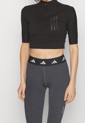 Kobieta w czarnym dopasowanym topie na zamek krótki rękaw oraz ciemnoszarych legginsach z wysokim stanem, z logo i markowym paskiem w talii.