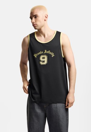 Młody mężczyzna z farbowanymi blond włosami nosi czarną bezrękawnik z napisem „Prosto Infinity 9” oraz luźne ciemne dżinsy, stoi na białym tle.