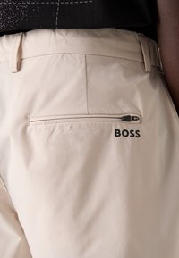 Beiga byxor har en slät yta, en bakficka med dragkedja och en svart "BOSS"-logotyp. Midjan är skräddarsydd och sitter bekvämt.
