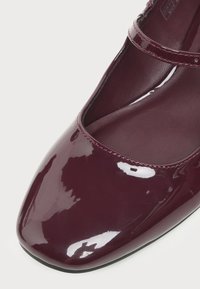 Chaussure Mary Jane bordeaux foncé brillant avec un bout carré arrondi et une fine bride sur le cou-de-pied sur fond blanc.