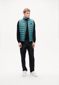 Gilet rembourré bleu clair avec des coutures horizontales, t-shirt noir à manches longues, pantalon en denim foncé et sneakers blanches. Design minimaliste, sans motifs.