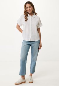 Biała lniana koszula z krótkimi podwiniętymi rękawami, z zapięciem na guziki, zestawiona z jasnoniebieskimi dżinsami i białymi sneakersami z brązowymi podeszwami.