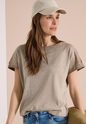 Kvinde iført en beige kasket og løs beige t-shirt kombineret med blå jeans, der holder en lysebrun jakke mod en lys baggrund.