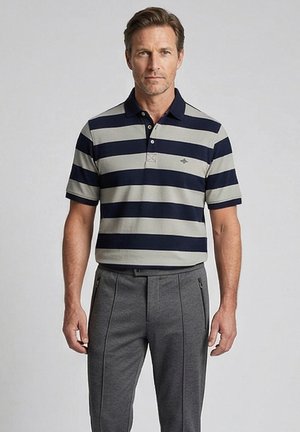 Man die een navyblauw en grijs horizontaal gestreept poloshirt en grijze broek draagt, staand tegen een effen lichte achtergrond.