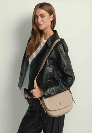 Młoda kobieta o długich brązowych włosach nosi czarną skórzaną kurtkę i trzyma na ramieniu beżową torbę przez ramię, delikatnie się uśmiechając.