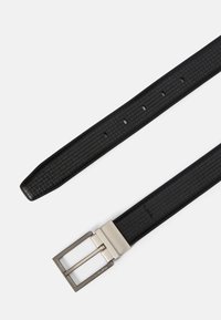 Μαύρη δερμάτινη ζώνη με υφή, πλεχτό σχέδιο και ορθογώνια ασημένια μεταλλική πόρπη. Περιέχει αρκετές τρύπες ρύθμισης κατά μήκος της ζώνης.