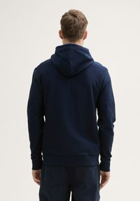 Hanorac bleumarin dintr-un material texturat, cu glugă reglabilă, manșete ribbate și o talie ajustată, prezentat din spate.