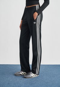 Čierne športové nohavice s bielymi bočnými pruhmi, so šnúrkou v páse a vreckami na zips. Nosia sa so sivými a bielymi športovými topánkami.