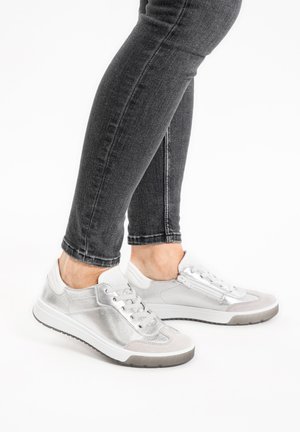 Person wearing silver lace-up sneakers with white soles and black skinny jeans, standing on a white background.