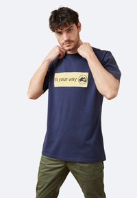 Camiseta de algodón azul marino de manga corta con un rectángulo gráfico amarillo que dice "es tu camino" y un logo. Textura suave.