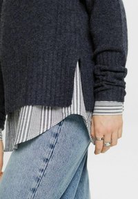 Dunkelgrauer Strickpullover mit geripptem Muster, über einem grau-weiß gestreiften Hemd getragen. Legere blaue Jeans aus Denim mit leichtem Verblassen.