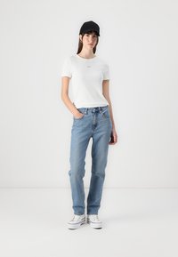 Valge lühikeste varrukatega T-särk logoga, helesinised sirged teksad ja must müts. Mudel kannab valgeid sneaker'eid, käed taskutes.