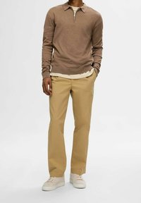 Pull en maille marron clair avec un col zippé, superposé à une chemise crème, associé à un pantalon beige et des baskets blanches, affichant une coupe décontractée.