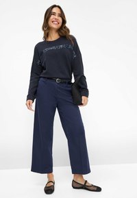 Marineblauw sweatshirt met lange mouwen en pailletten, gecombineerd met marineblauwe broek met wijd uitlopende pijpen en zwarte mesh ballerina's, aangevuld met een zwarte tas.