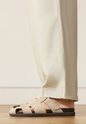 Beige gewebte Stoffsandalen mit verstellbaren Riemen, getragen zu hellbeigen Hosen, seitlich auf einem Holzboden gezeigt.