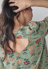 Blusa verde con estampado floral que presenta mangas abullonadas, una espalda recortada y flores multicolores en naranja, rojo y púrpura. Textura de tela suave.