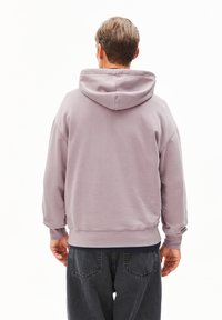 Uomo con un felpa viola chiaro con il cappuccio abbassato e jeans neri, in piedi con le spalle rivolte verso di noi su uno sfondo bianco.