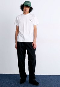 White short-sleeve t-shirt with a logo on the left chest, paired with black trousers and a green cap, standing on a blue carpet.