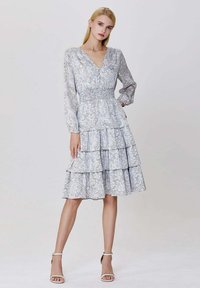 Femme aux cheveux blonds raides portant une robe midi à manches longues, à volants et à fleurs, et des talons ouverts blancs, se tenant devant un fond uni.