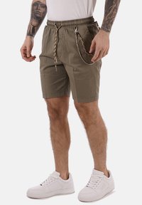 Pantaloni corte verde oliva con vita elastica, cordino e cerniere laterali. Indossati con sneakers bianche, caratterizzate da una texture liscia.