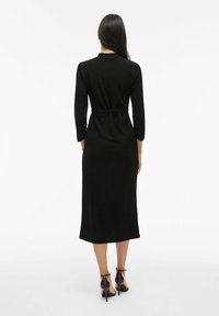 Black, mid-length dress with long sleeves, fitted waist, and a belt. Smooth fabric, simple design, and paired with burgundy heeled shoes.