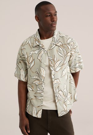 Muž v béžovej košeli na gombíky s veľkými bielymi a hnedými listovými vzormi priamo na svetlohnedom tričku.