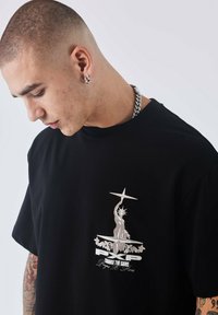 Zwarte katoenen t-shirt met een zilveren afbeelding van het Vrijheidsbeeld en de tekst "VERANDER HET SPEL" met decoratieve accenten.