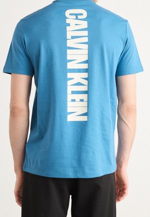 Personne portant un t-shirt bleu avec "CALVIN KLEIN" imprimé verticalement en blanc dans le dos, associé à un short noir.