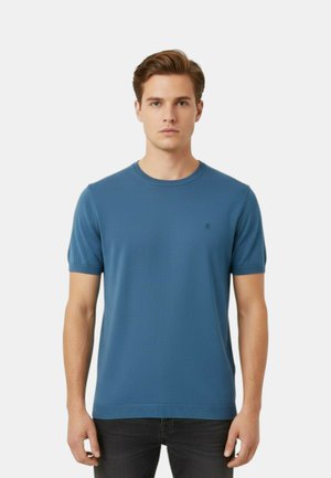 Giovane uomo che indossa una maglietta blu tinta unita a maniche corte in maglia e pantaloni neri, in piedi davanti a uno sfondo chiaro e neutro.