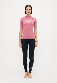 Maglietta a maniche corte rosa con logo a cuore bianco, abbinata a leggings neri con il nome del marchio "ROXY" in bianco su una gamba.