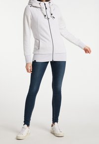 Světlá šedá mikina na zip s šňůrkami, má měkkou texturu a boční kapsy, v kombinaci s tmavě modrými skinny džíny a bílými teniskami.