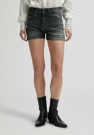 Kvinde iført sorte denimbukser, en hvid bluse med lange blonderærmer og sorte læderstøvler til midt på læggen, stående på en grå baggrund.