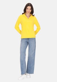 Langærmet gul pullover med kvart-zip krave, parret med lyseblå wide-leg jeans og hvide sneakers. Blød tekstur, afslappet design.