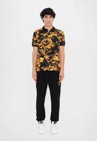 Camisa polo preta com padrões amarelos ornamentados, acompanhada de calças jogger pretas. Modelo usa ténis brancos. O fundo é branco liso.