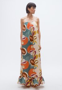 Γυναίκα που φοράει μακρύ αμάνικο φόρεμα με λαιμόκοψη halter και πολύχρωμο αφηρημένο λουλουδάτο σχέδιο και βολάν στο τελείωμα, όρθια ξυπόλητη, κοιτάζοντας προς τα πλάγια.