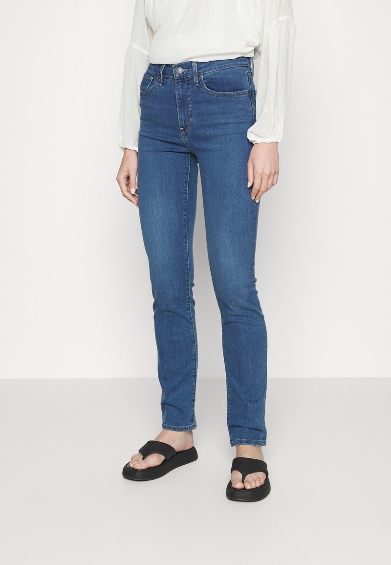Femme portant un jean bleu slim, un chemisier blanc à manches longues et des sandales noires à bouts ouverts, debout devant un fond clair uni.
