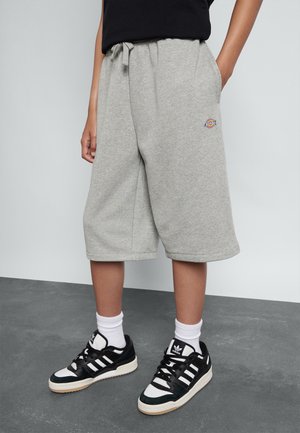 Osoba w szarych, do kolan sięgających spodenkach z małym logo, czarno-białych butach Adidas, białych skarpetkach i czarnej, wsuniętej koszulce.