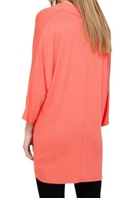 Tunique oversize corail à manches trois-quarts, coupe décontractée et texture lisse, descendant jusqu'à mi-cuisse. Design minimaliste.