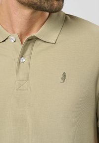 Beige polo paita, joka on valmistettu teksturoidusta kankaasta ja jossa on kaksi nappia, pieni brodeerattu hahmo vasemmassa rinnassa sekä perinteinen kauluskanta.