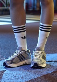 Sportovní obuv v šedé a bílé síťovině s černými akcenty a třemi pruhy. V kombinaci s bílými žebrovanými ponožkami s černými pruhy a logem.