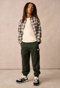 Child wearing a green cargo pants, a cream graphic t-shirt, and a black and gray plaid jacket, paired with black and white sneakers.