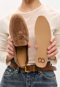 Mocasín de ante marrón con un acento metálico, que presenta detalles de costura y una suela beige lisa sostenida junto al zapato.