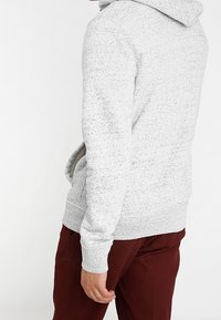 Sudadera gris claro hecha de una tela suave y texturada, con un bolsillo frontal. Presenta puños y dobladillo acanalados, con un corte holgado.