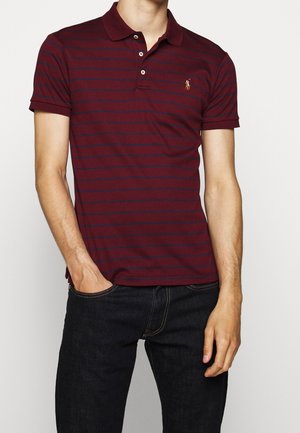 Burgunderfarbenes Polohemd mit blauen Streifen, kurzen Ärmeln und einem Kragen, versehen mit einem kleinen gestickten Logo auf der linken Brust. Getragen mit dunkler Jeans.