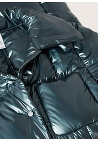 Polstret pufferjakke i dyb mørk blågrøn farve, med et blankt, reflekterende materiale, trykknapper og quiltede sektioner for ekstra tekstur.